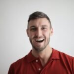 A cheerful man smiling broadly in a red polo shirt against a white background.